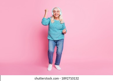 Full length photo of pensioner lady dance raise fist wear blue sweater denim jeans earphones sneakers isolated pink color background
