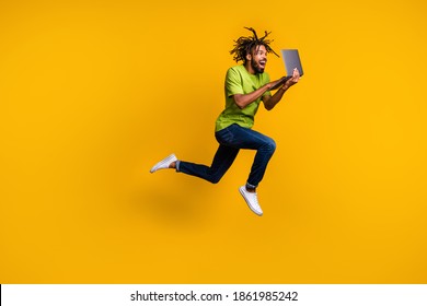 Full length photo portrait of excited programmer with dreadlocks jumping up holding laptop in hands isolated on vivid yellow colored background
