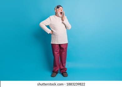 Full Length Photo Portrait Of Yawning Old Man Covering Mouth With Hand In Eye Mask Isolated On Pastel Blue Colored Background