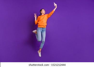 Full Length Photo Of Positive Cheerful Youth Teen Girl With  Ponytails Have Trip Journey Take Selfie Make V-signs Wear Orange Sportive Wear Jeans Sneakers Isolated Violet Purple Color Background