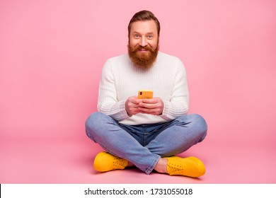 Full length photo of positive cheerful man sit floor legs crossed use cell phone read social media news texting typing sms wear jeans jumper trendy shoes isolated pastel color background