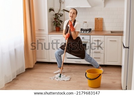 Full length photo of positive cheerful woman washing floor imagine she real celebrity holding mop, singing song, wear latex gloves and brown apron, posing in house indoors.
