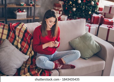 Full length photo of positive girl sit couch text cellphone wear red sweater in house indoors with x-mas christmas decoration