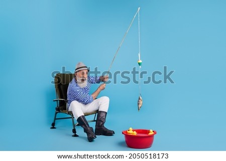 Full length photo of positive happy grey haired old man smile catch fish sit chair vacation isolated on blue color background