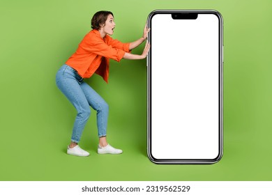 Full length photo of positive impressed lady dressed jeans shirt holding modern gadget empty space isolated green color background