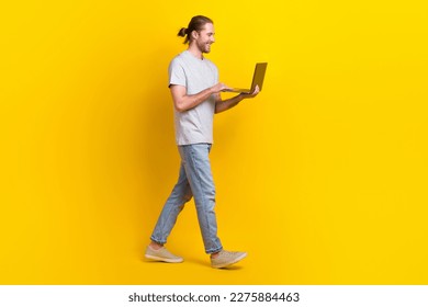 Full length photo of positive satisfied guy wear light t-shirt jeans holding laptop walk empty space isolated on yellow color background