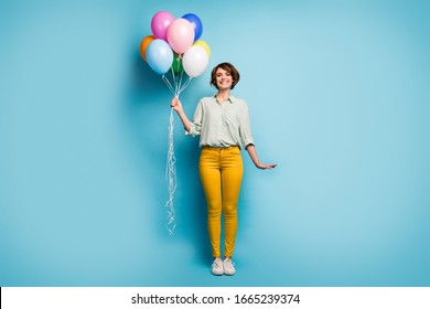 Full length photo of pretty cute adorable lady surprise birthday party hold many colorful air balloons wear casual green shirt yellow pants shoes isolated blue color background - Powered by Shutterstock