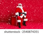 Full length photo of retired old man white beard sit big giftbox hold dumbbells warming up arms wear x-mas santa costume glove coat belt sunglass cap boot isolated red color background