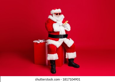 Full length photo of retired old man white beard hold telephone sit large box what chatting elf friend wear x-mas santa costume sunglass headwear leather boot isolated red color background - Powered by Shutterstock