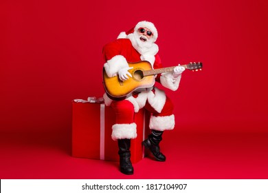 Full length photo of retired old man white beard hold guitar sit box play sing christmas carol song wear santa x-mas costume sunglass headwear leather boot isolated red color background - Powered by Shutterstock