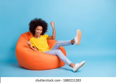 Full length photo of small dark skin happy girl celebrate win sit bean ban wear yellow t-shirt isolated on blue color background - Shutterstock ID 1880211283