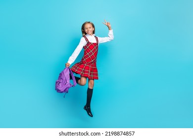 Full length photo of small schoolgirl jump hold bag demonstrate v-sign isolated on blue color background