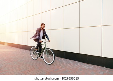 Full length photo of smiling young bearded man riding a bike on the city street. Dressed up in plaid shirt, t-shirt and jeans. Recreation concept. - Powered by Shutterstock