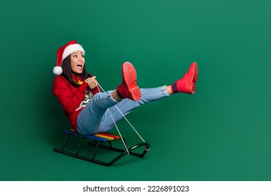Full length photo of sweet funky girl dressed ugly print red x-mas pullover riding sledge looking empty space isolated green color background