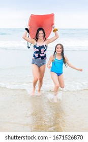 Full Length Photo Of Two Cute Teen Girls Running On A Beautiful Beach Carrying A Bodyboard. Having Fun In The Sun While On A Family Beach Vacation.	