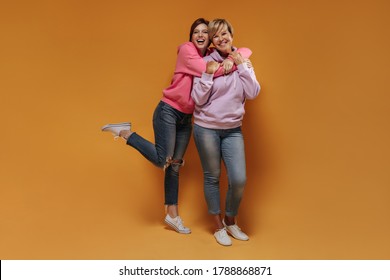 Full Length Photo Of Two Smiling Women With Short Hairstyle In Cool Hoodies, Skinny Jeans And Light Sneakers Hugging On Orange Backdrop..