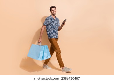 Full length photo of walking businessman carry brand new clothes shopaholic hold phone got promocode isolated on beige color background