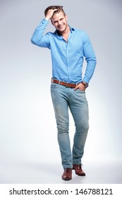 full length photo of a young casual  man standing with a hand in his pocket and the other in his hair while smiling for the camera. on gray studio background