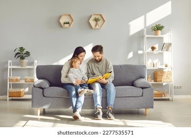 Full length photo of a young happy family reading book with their little cute daughter sitting on the sofa at home. Parents spending time with their child and smiling. Leisure time together concept. - Powered by Shutterstock