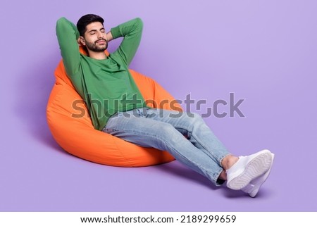 Full length photo of young latin man laying beanbag daydreaming chilling dressed stylish green garment isolated on purple color background