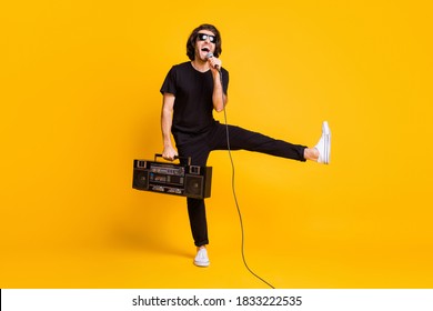 Full Length Photo Of Young Man Hold Vintage Radio Mic Open Mouth Raise Leg Wear Black T-shirt Pants White Sneakers Glasses Isolated Yellow Color Background