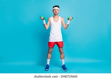 Full length photo of young man lift who dumbbells biceps training make funny face heavy isolated on blue color background