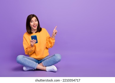 Full length photo of young pretty girl sitting using new smartphone blogger finger directing empty space eshopping offer isolated on violet color background