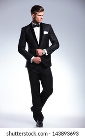 full length picture of an elegant young fashion man adjusting his tuxedo while looking to his side, away from the camera. on gray background