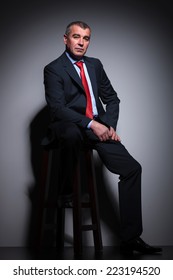 Full Length Picture Of A Mid Aged Business Man Resting On A Stool, Looking At The Camera.