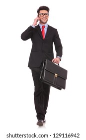 Full Length Picture Of A Young Business Man Walking Forward With A Briefcase In One Of His Hands And Talking At The Phone While Looking Away From The Camera On White Background