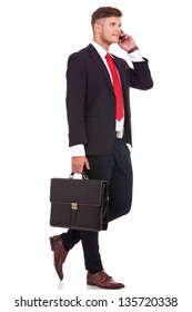 Full Length Picture Of A Young Business Man Walking With His Briefcase And Talking On The Phone While Looking Away From The Camera. Isolated On White Background