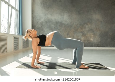 Full Length Picture Of Young Pregnant Caucasian Yoga Couch Showing Exercise On Black Mat, Face Turned To Ceiling, Straight Hands On Mat, Legs Bend In Knees, Back Facing Floor For Reaching Balance