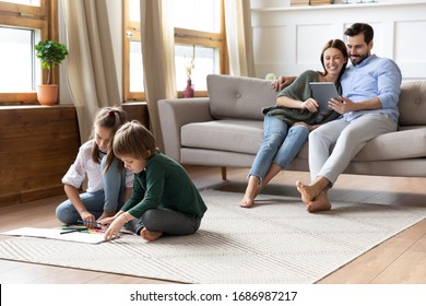 Full Length Playful Little Children Siblings Sitting On Floor Carpet Drawing Pictures While Addicted To Technology Smiling Parents Resting On Couch With Computer Tablet, Family Weekend Pastime.