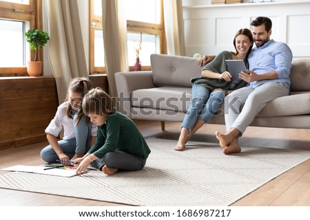 Full length playful little children siblings sitting on floor carpet drawing pictures while addicted to technology smiling parents resting on couch with computer tablet, family weekend pastime.