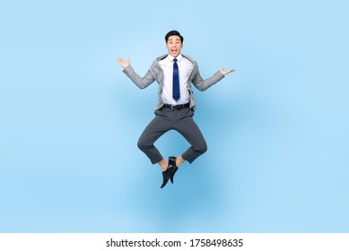 Full length playful portrait of happy ecstatic young Asian businessman jumping in mid-air doing wacky fun gesture in isolated studio blue background