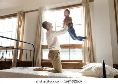 Full length playful young father standing on knees on comfortable mattress bed, lifting little preschool daughter in air. Happy small child girl flying, having fun with joyful single daddy in bedroom.