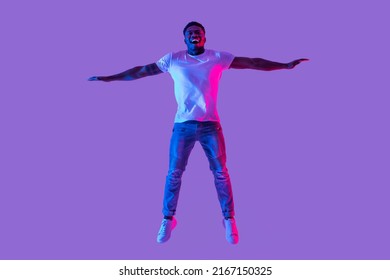 Full length portrait of active energetic young black man jumping in neon light, copy space. Excited African American guy celebrating success or victory, flying in air, expressing joy and happiness