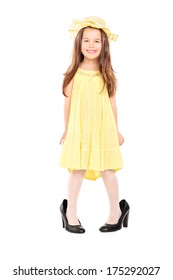 Full Length Portrait Of Adorable Little Girl In Fancy Yellow Dress And Hat Standing In Oversized High Heels Isolated On White Background