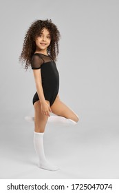Full length portrait of adorable smiling girl in black sportswear and white knee socks posing with leg bent, isolated on gray studio background. Little female professional gymnast with curly hair.