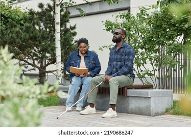 Full Length Portrait Of Adult Couple With Partner With Visual Disability Sitting On Bench In Park Together