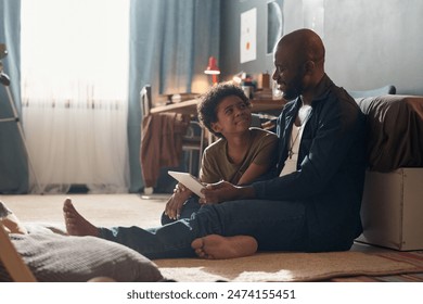 Full length portrait of African American single father and smiling son using tablet and chatting sitting on floor in harsh sunlight copy space - Powered by Shutterstock