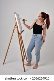 Full Length Portrait Of Artist Girl Wearing Basic White Shirt And Jeans. Standing Pose  Holding A Paintbrush And Palette, Painting On A Black Canvas Easel.   Isolated On  Studio Background 