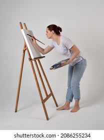 Full Length Portrait Of Artist Girl Wearing Basic White Shirt And Jeans. Standing Pose  Holding A Paintbrush And Palette, Painting On A Black Canvas Easel.   Isolated On  Studio Background 