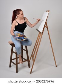 Full Length Portrait Of Artist Girl Wearing Basic White Shirt And Jeans. Sitting Pose  Holding A Paintbrush And Palette, Painting On A Black Canvas Easel.   Isolated On  Studio Background 