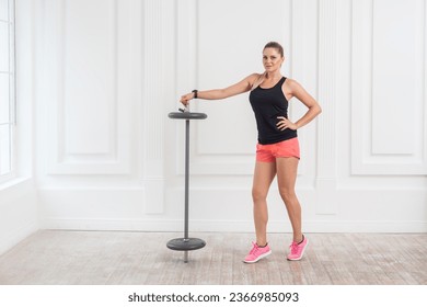 Full length portrait of athletic beautiful woman in pink top and black shorts standing, holding barbell and looking at camera with confident expression on white wall background. Indoor studio shot. - Powered by Shutterstock