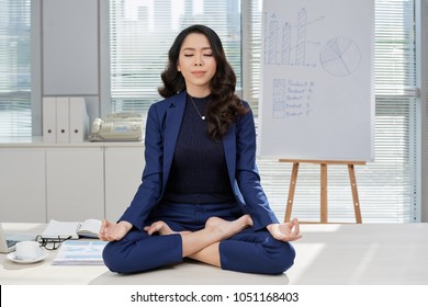 Full length portrait of attractive Asian businesswoman sitting on office desk in lotus position while doing yoga in order to relieve stress - Powered by Shutterstock