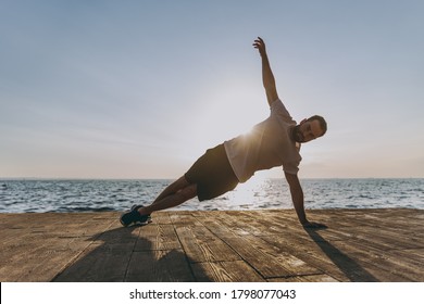 Full length of portrait of attractive handsome young bearded athletic man 20s in casual white t-shirt posing training doing side plank exercise looking camera at sunrise over the sea outdoors