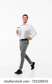 Full Length Portrait Of An Attractive Joyful Man In A White Shirt Holding Laptop Computer And Coffee Cup While Walking And Looking At Camera Isolated Over White Background