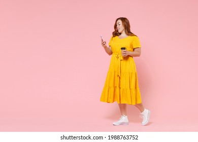 Full length portrait attractive young redhead plus size body positive female woman 20s in yellow dress hold paper cup coffee or tea using mobile phone isolated on pastel pink color background studio - Powered by Shutterstock