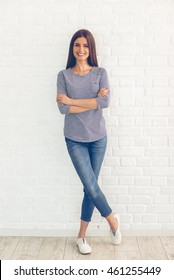 Full length portrait of attractive young woman in casual clothes looking at camera and smiling while standing with crossed arms in front of white wall